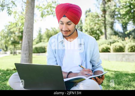 Ein indischer Student mit rotem, traditionellem Turban-Pheta, der mit einem Laptop online studiert, auf dem Rasen sitzt, sich ein Webinar ansieht und sich Notizen macht, um sich auf die Prüfungen in der Nähe des Campus im Freien vorzubereiten Stockfoto