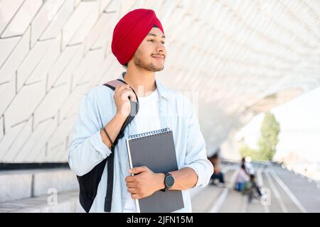 Inspirierter indischer Student mit Rucksack, der Laptop im Freien hält, Ausländer hindu, der stolz darauf ist, Universitätsmitglied zu sein, hindu im traditionellen Turban, der im Freien fröhlich zur Seite steht Stockfoto