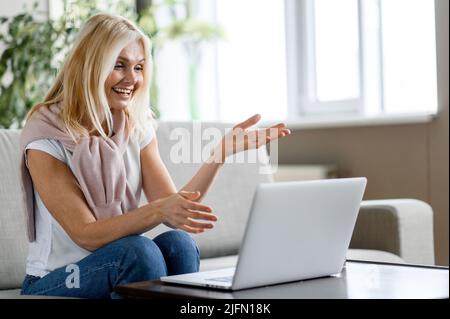 Fröhliche, freundliche, kaukasische blonde Frau mittleren Alters in legerer Kleidung, sitzt auf einem gemütlichen Sofa im Wohnzimmer, spricht auf einer Videokonferenz mit Familie, Freunden oder Kollegen, mit einem Laptop, lächelt Stockfoto