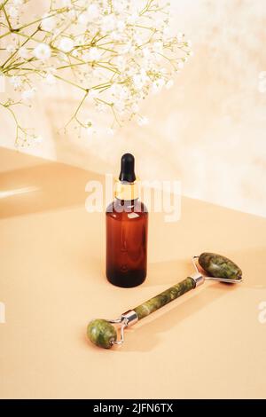 Serumflasche und Jade-Gesichtswalze auf neutralem beigen Hintergrund mit Gypsophila-Blüten, Nahaufnahme Stockfoto