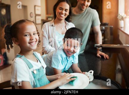 Junge glückliche Mischling-Eltern waschen das Geschirr mit ihrem Sohn und ihrer Tochter in der Küche zu Hause. Kleiner Bruder und Schwester helfen ihrer Mutter und Stockfoto