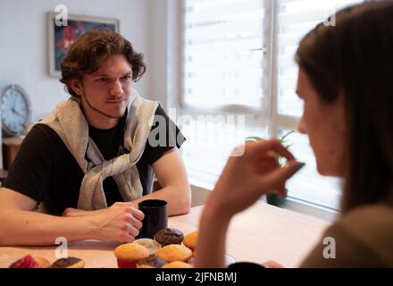 Junger Mann und Frau sitzen am Tisch, trinken Kaffee und essen Muffins zu Hause Stockfoto
