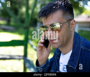 Ein hübscher Mann in einer Jeansjacke, der im Park eine Sonnenbrille trägt und mit dem Handy spricht Stockfoto