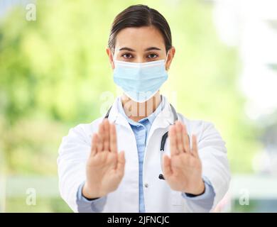 Porträt einer hispanischen Ärztin, die eine Maske trägt und mit ihrer Hand eine Stop-Geste zeigt, während sie in einem Krankenhausbüro ein Stethoskop trägt. Stockfoto