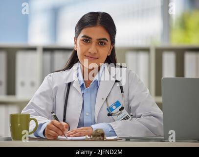 Porträt einer jungen indischen Ärztin mit einem Stethoskop, die in einem Büro sitzt und ein Rezept schreibt, während sie an ihrem Schreibtisch sitzt. Gemischter GP mit einem Stockfoto