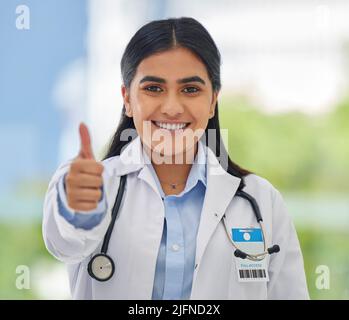 Portraitaufnahme eines jungen glücklichen Mixed Race Doktors, der sich einen Daumen zeigt, während er allein in einem Krankenhaus arbeitet. Ein glücklicher, hispanischer gp lächelte und zeigte Stockfoto