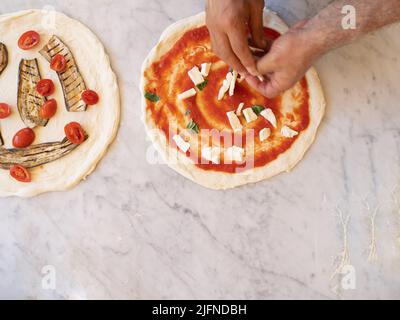 Foto des traditionellen weißen Pizzateigs mit Platz zum Kopieren auf einer Marmorplatte, Koch fügt Mozzarella-Käse ein Basilikum hinzu, er bereitet sich darauf vor, gebacken zu werden Stockfoto