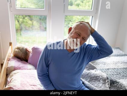 Älterer Mann sitzt auf dem Bett und streckt sich morgens den Hals, während die Frau noch im Hintergrund schläft Stockfoto