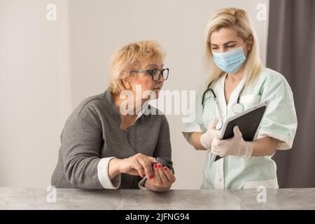 Hausgesundheitspflege. Frau Krankenschwester in einer medizinischen Maske helfen mittleren Alters Frau während der Krankheit oder Druck, weibliche Ärztin misst den Puls des Patienten Stockfoto