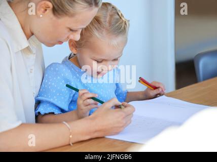 Mutter unterrichtet kleine Tochter während der Homeschool-Klasse zu Hause. Nettes kleines kaukasisches Mädchen, das lesen und schreiben lernt, während ihr Elternteil ihr hilft Stockfoto