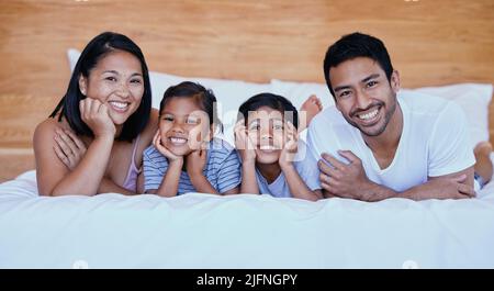 Glückliche Familie im Bett. Porträt einer jungen hispanischen Familie im Bett. Junge gemischte Rasse Familie von vier. Zwei Eltern, die sich zu Hause mit ihren Töchtern verbinden Stockfoto