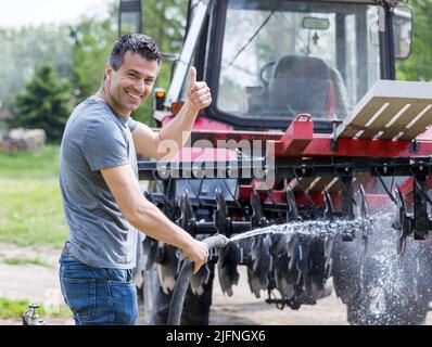 Schöner zufriedener Mann, der Traktor und Ausrüstung mit Wasserstrahl aus dem Schlauch putzt und Daumen auf dem Bauernhof zeigt Stockfoto