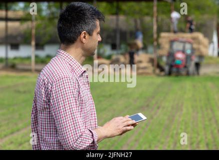 Porträt eines gutaussehenden Bauern in schlichtem Hemd, der auf dem Bauernhof mit Traktor und Arbeitern im Hintergrund auf dem Tablet arbeitet Stockfoto