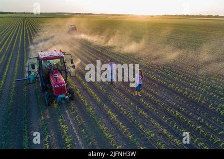 Luftbild eines älteren Bauern, der im Maisfeld steht, und einer Geschäftsfrau, die beim Schlagen von Boden durch den Traktor von einer Drohne aus schießt Stockfoto