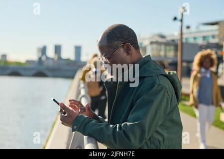 Afrikanischer reifer Mann in einer Brille, der eine Nachricht auf seinem Mobiltelefon tippt, das im Freien in der Stadt steht Stockfoto