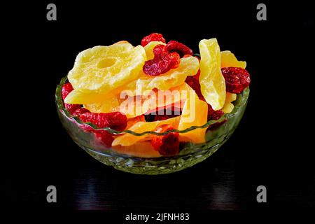 Gelbe kandierte Melonenscheiben, Ananasringe und Dogwood in einer Glasvase auf schwarzem Hintergrund. Stockfoto