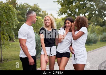 Selbstbewusster, gutaussehender Mann, der im öffentlichen Park mit drei jungen, sportlichen Frauen flirtet, asiatische und schwarze Damen, die ihn diskutieren und lachen Stockfoto