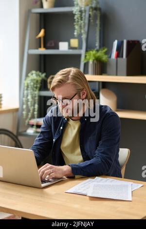 Junger Geschäftsmann in einer Brille, der am Laptop tippt und an seinem Arbeitsplatz im Büro online arbeitet Stockfoto