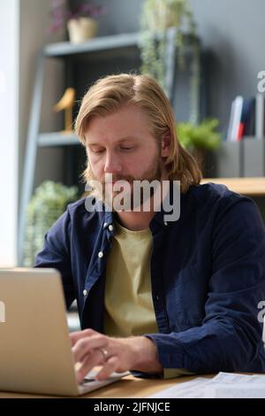 Junger bärtiger Büroangestellter, der in zwangloser Atmosphäre einen Laptop für Online-Arbeiten am Schreibtisch verwendet Stockfoto