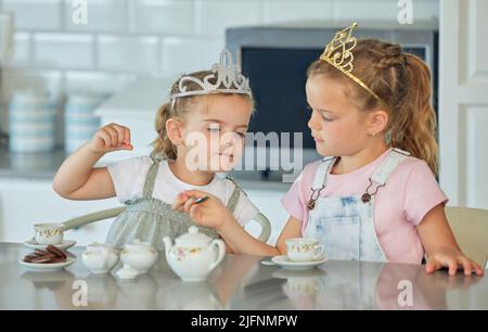 Zwei kleine Mädchen mit einer Prinzessin Tee-Party zu Hause. Geschwister oder Freunde tragen Tiaras, während sie mit dem Teeset spielen und in der Küche Kekse trinken Stockfoto