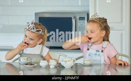 Zwei kleine Mädchen mit einer Prinzessin Tee-Party zu Hause. Geschwisterfreunde tragen Tiaras, während sie mit dem Teeset spielen und in der Küche Kekse trinken Stockfoto