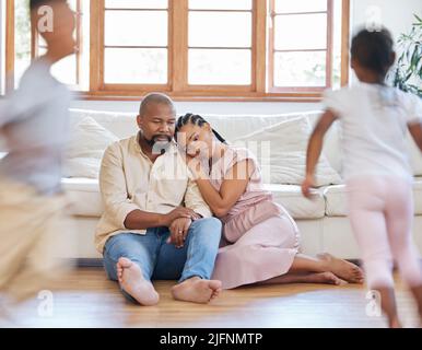 Müde afroamerikanische Paare, die auf dem Loungefußboden sitzen und müde und erschöpft aussehen, während ihre Hyper Kinder um sie herum spielen. Zwei schwarze Geschwister Stockfoto