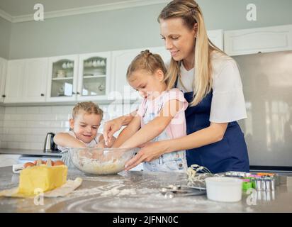 Lächelnde kaukasische Mutter mit zwei entzückenden Töchtern, die zu Hause backen. Mama und Kinder mischen Teig oder Zutaten in einer Schüssel für Pfannkuchen oder Teig für Stockfoto