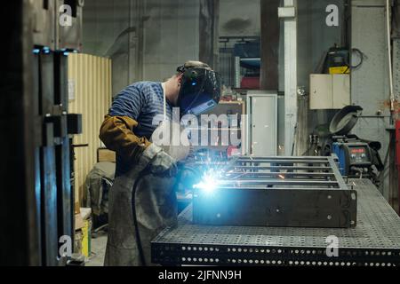 Seitenansicht des männlichen Arbeiters einer riesigen Industrieanlage oder Manufaktur, der sich während der Schweißarbeiten über eine große metallische Werkbank biegt Stockfoto