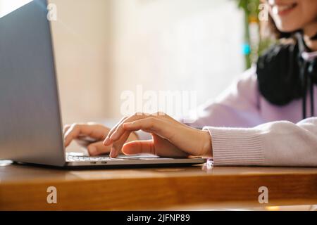 Junge asiatische Frau in Kopfhörern lächelt und tippt auf dem Laptop im Café Stockfoto