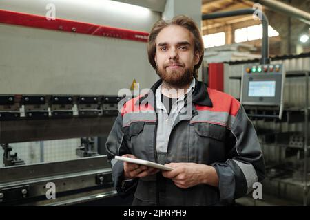 Junge selbstbewusste Ingenieur mit Tablet vor der Kamera in der Werkstatt der Industriefabrik stehen und im Netz arbeiten Stockfoto