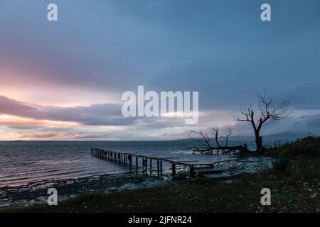 Langzeitansicht eines Piers auf einem See in der Dämmerung, mit vollkommen stillem Wasser, sich bewegenden Wolken und Skelettbäumen Stockfoto