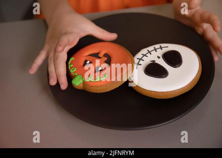 Die Hand des Mädchens greift nach hausgemachten Halloween-Keksen mit bemalten Gesichtern Stockfoto