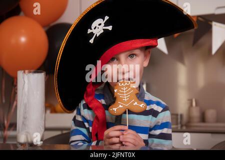 Ein als Pirat gekleideter Junge isst Lebkuchen als Halloween-Skelett, trinkt Milch Stockfoto