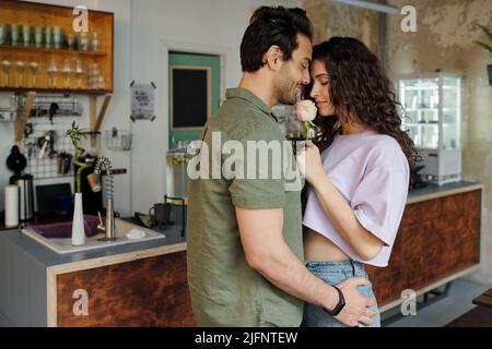 Junge liebevolle Mann in der Nähe seiner wunderschönen Freundin riechende rosa Rose, nachdem sie ihre Überraschung während der romantischen Date im Café Stockfoto