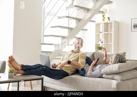 Glückliches reifes älteres Paar, das mit Geräten spricht, die sich zu Hause auf der Couch entspannen. Stockfoto