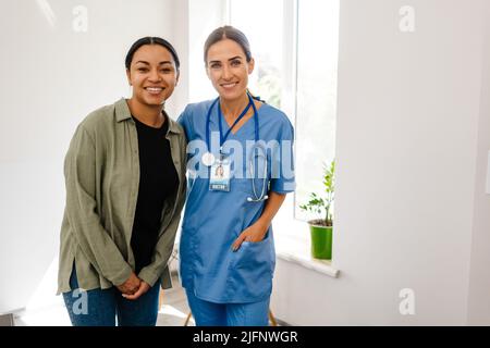 Weiße Ärztin lächelt, während sie mit ihrem Patienten im Büro im Haus posiert Stockfoto