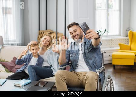 Kaukasischer Mann mit Behinderungen hält Smartphone und nimmt Selfie mit der Familie zu Hause. Lächelnde Mutter und Sohn sitzen zusammen auf dem Sofa in der Nähe des Vaters im Rollstuhl und winken die Hände auf der Telefonkamera. Stockfoto