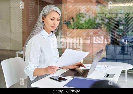 Seriöse Geschäftsfrau mittleren Alters, die Papierkram mit pc-Laptop-Computer macht. Stockfoto