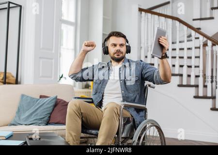 Ein hübscher Rollstuhlfahrer mit Kopfhörern tanzt mit den Händen zu netter Musik in einer modernen Wohnung. Bärtiger Mann mit Behinderung hält digitale Tablette für die Wahl neuer Spur. Stockfoto