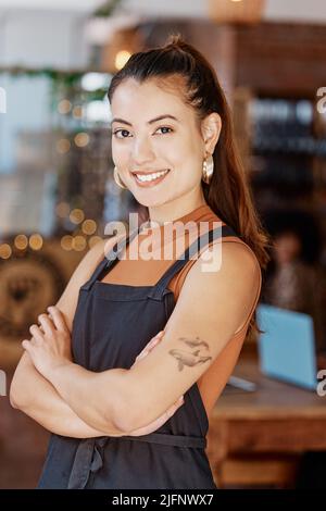 Porträt einer lächelnden, schönen asiatischen Geschäftsbesitzerin, die mit gekreuzten Armen in ihrem Café steht. Selbstbewusste und erfolgreiche Mixed-Race-Frau trägt Stockfoto