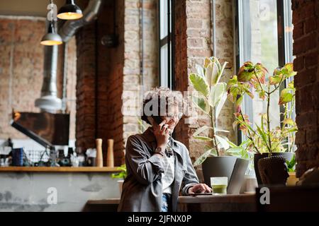 Glückliche junge Besitzerin eines modernen Cafés, die Kunden am Telefon und online berät, während sie vor dem Laptop an einem großen Fenster sitzt Stockfoto