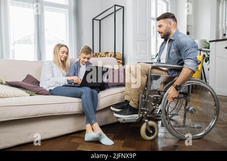 Behinderter Mann im Rollstuhl, der mit seiner lieben Frau und seinem hübschen Sohn im Schulalter spricht und zu Hause einen digitalen Laptop verwendet. Junge Frau und Kind sitzen auf der bequemen Couch. Stockfoto