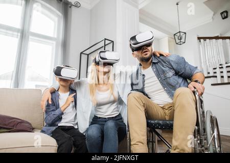 Attraktive Mutter umarmt niedlichen Sohn und bärtigen Vater im Rollstuhl, während mit vr Headset in gemütlichen Hause. Konzept von glücklicher Familie, moderner Technik und Freizeit. Stockfoto