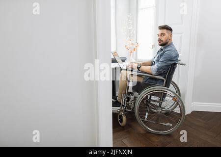 Fokussierter bärtiger Mann, der im Rollstuhl einen Laptop auf den Knien hält und die Kamera anschaut. Fleißiger freier männlicher Freiberufler mit Behinderung, der von zu Hause aus arbeitet. Stockfoto