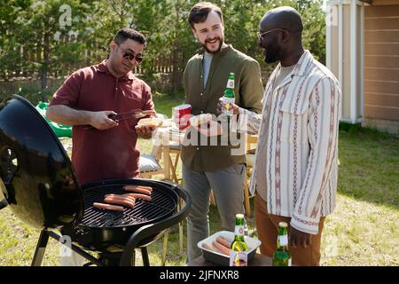 Drei glückliche junge interkulturelle Männer, die am Wochenende Bier trinken und sich während einer Freiluftparty unterhalten, während einer von ihnen grillen lässt Stockfoto