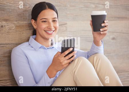 Junge glückliche Geschäftsfrau mit gemischtem Rennen nutzt soziale Medien am Telefon, während sie während einer Pause auf dem Boden bei der Arbeit eine Tasse Kaffee trinkt. Gemischt Stockfoto