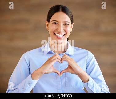 Junge glückliche hispanische Geschäftsfrau lächelt und steht und macht eine Herzgeste mit ihren Händen allein bei der Arbeit. Fröhliche und positive gemischte Rasse Stockfoto
