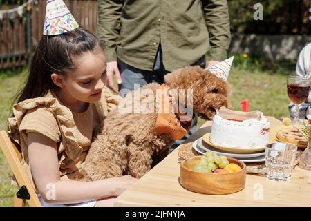 Netter Haustier Hund von einem kleinen Mädchen gehalten, das Geburtstagskuchen mit Knochen isst, der während des Abendessens im Freien oder einer Hinterhofparty auf dem Tisch steht Stockfoto