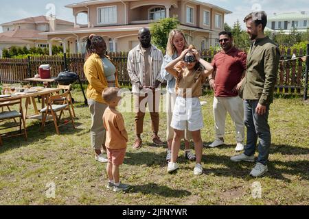 Junge Frau blindlinding kleine Tochter beim Spielen verstecken und suchen mit Freunden auf grünem Gras des Hinterhofs des Landhauses Stockfoto