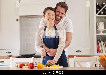 Wir stecken Liebe in das, was wir tun. Aufnahme eines Mannes, der seine Frau umarmt, während sie in der Küche eine Mahlzeit zubereitet. Stockfoto